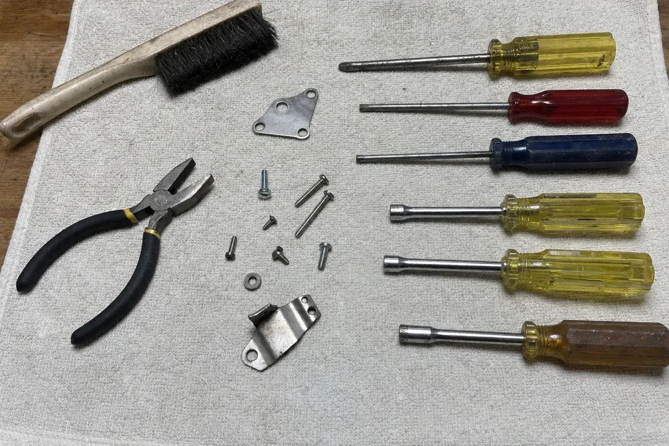 Hand tools laid out on canvas — wire brush, pliers, six drivers, loose screws and two sheet-metal brackets